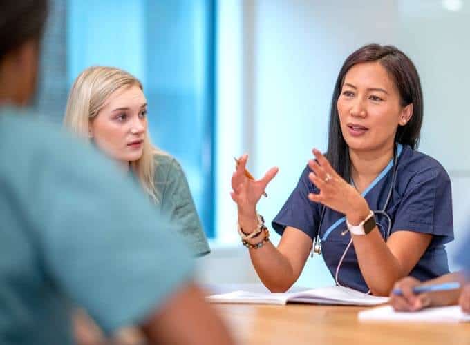 A small group of Nurse sit at a boardroom table as they meet to discuss patient cases. They are each dressed professionally in scrubs and have stethoscopes around their necks as they discuss plans of care for each case.
